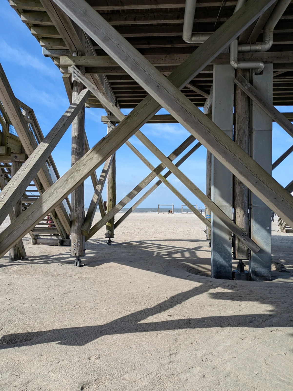 Blick unter einen Pfahlbau in St. Peter-Ording auf hohe Holzpfähle, gekreuzte Streben und den Sandboden