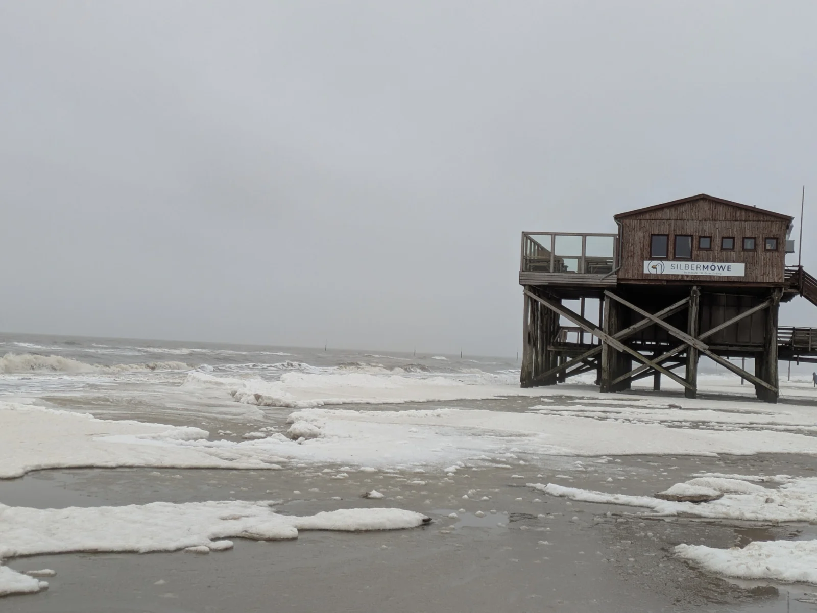 Hölzerner Pfahlbau mit Balkon und gekreuzten Stützen neben schäumenden Nordseewellen unter grauem Himmel in St. Peter-Ording