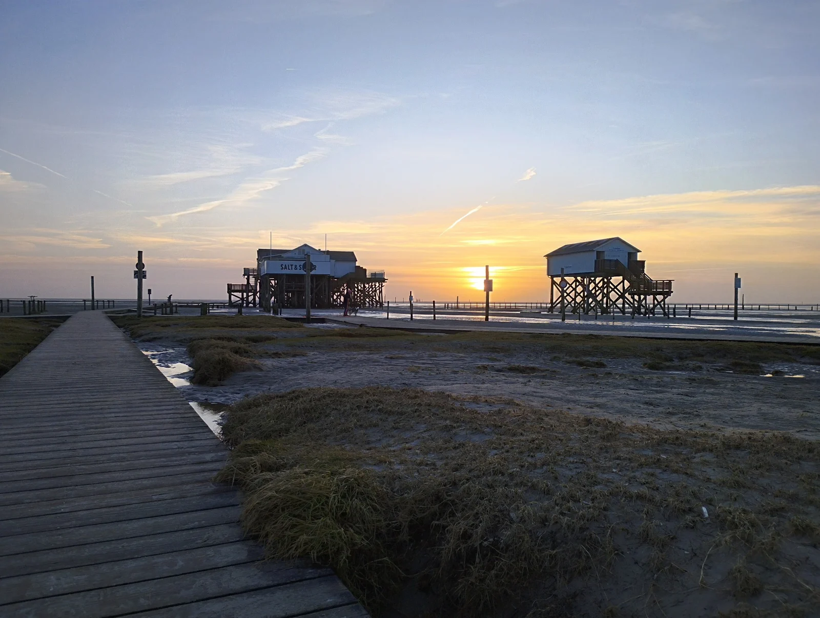 Zwei Pfahlbauten in St. Peter-Ording im goldenen Sonnenuntergang mit Holzsteg und Küstenwiesen im Vordergrund